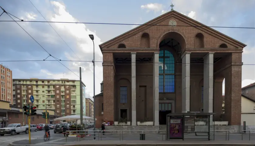 La facciata in mattoni rossi di una chiesa a Milano, con un grande rosone blu e colonne imponenti. Sullo sfondo, palazzi residenziali e tram.