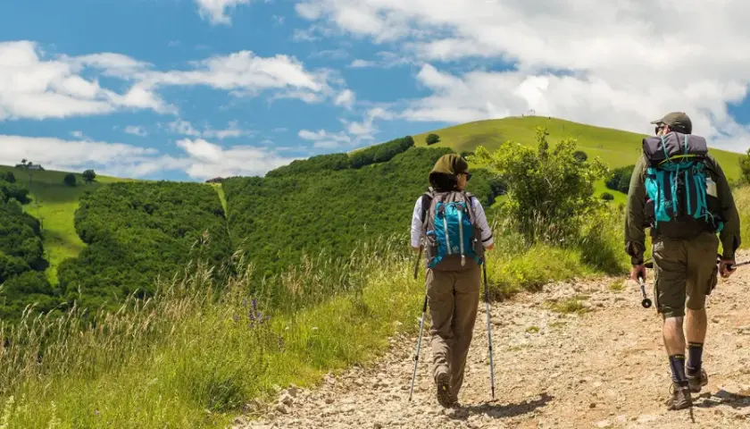 Due escursionisti affrontano un sentiero collinare, con lo sguardo rivolto verso l'orizzonte. La distanza tra Assisi e Loreto sembra un viaggio spirituale e fisico.