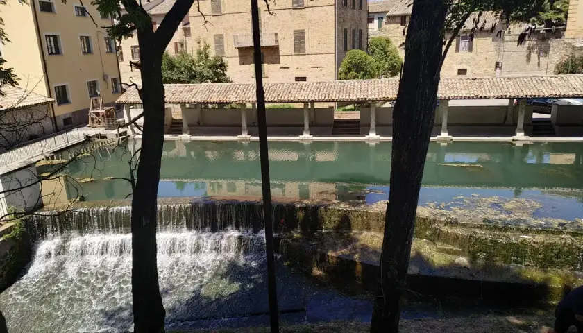 Cascata e portico storico a Bevagna, Umbria. L'acqua scende su gradoni di pietra, riflettendo gli edifici antichi.