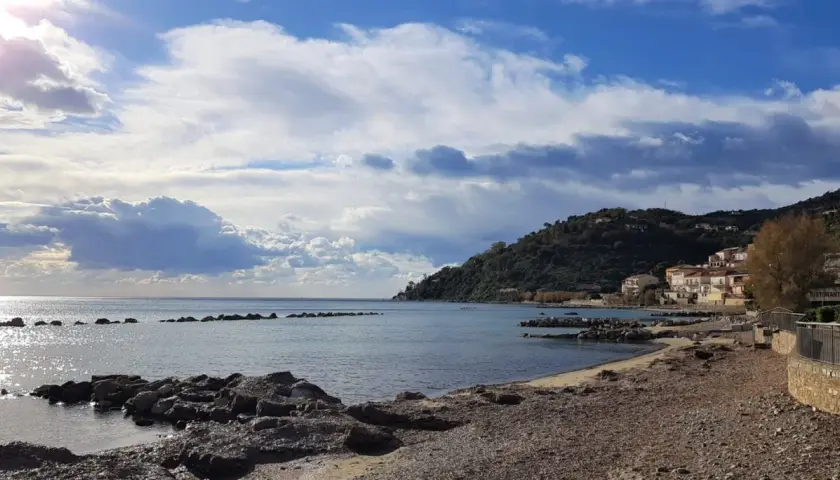 Costa di Pollica con case colorate arroccate sulla collina e mare calmo sotto un cielo nuvoloso.