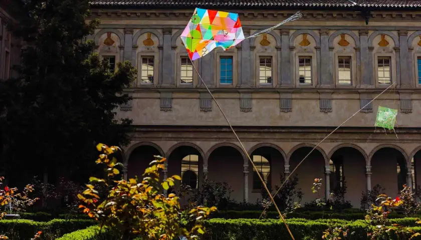 Coloratissimi aquiloni volano nel chiostro dell'Incoronata a Milano, tra siepi curate e un antico porticato.
