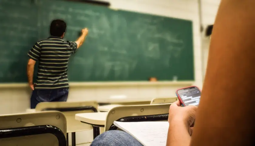 Studente distratto al cellulare mentre il professore scrive alla lavagna, forse un nuovo concorso scuola in arrivo.