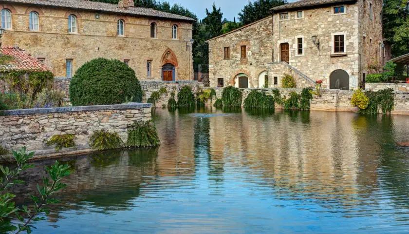 Antiche terme libere a San Gimignano, con edifici in pietra che si specchiano in un laghetto tranquillo.