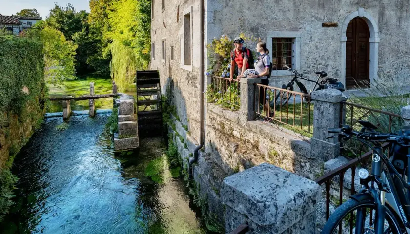 Coppia in bicicletta ammira un antico mulino ad acqua, uno dei tanti borghi friulani da visitare, immersi nel verde e lungo un ruscello cristallino.