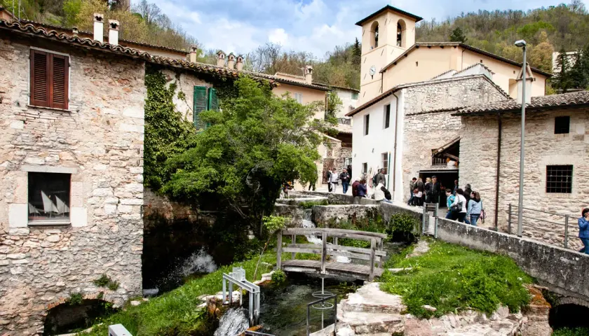 Un borgo in Umbria con cascate che scendono tra case di pietra e un ponte di legno.