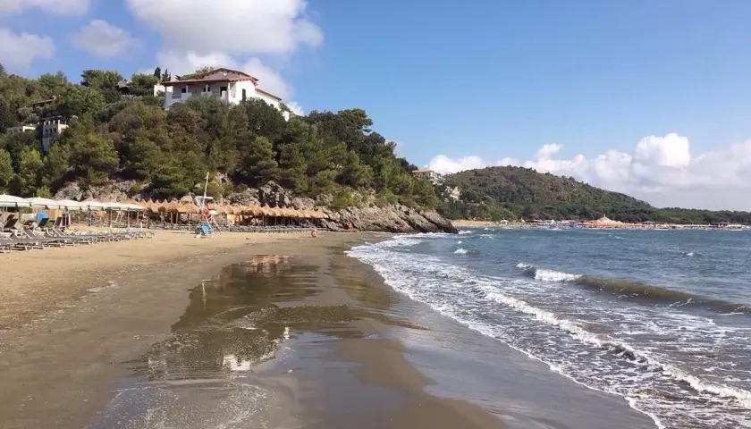 Spiaggia di **Porto d'Ercole** con ombrelloni, lettini e mare calmo sotto un cielo azzurro con nuvole bianche.