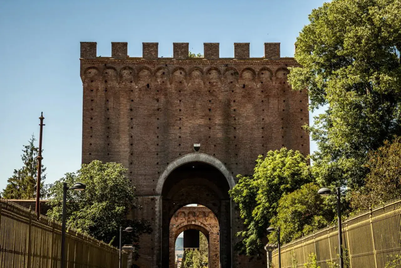 Imponente porta romana in mattoni con archi sovrapposti, circondata da alberi verdi e una recinzione dorata.