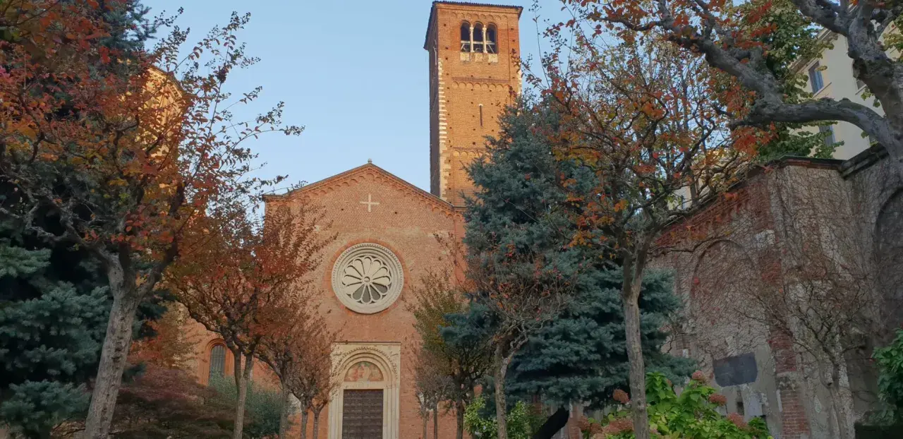 San Celso a Milano, con il suo campanile in mattoni rossi e la facciata ornata da un rosone, circondato da alberi dalle foglie autunnali.