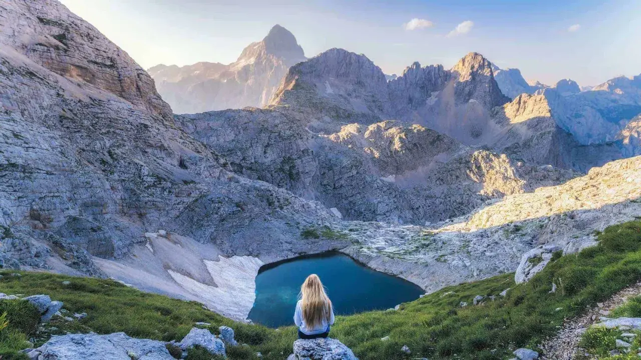 Donna con lunghi capelli biondi ammira un lago alpino nel Parco Nazionale del Tricorno, circondato da maestose vette rocciose illuminate dal sole.