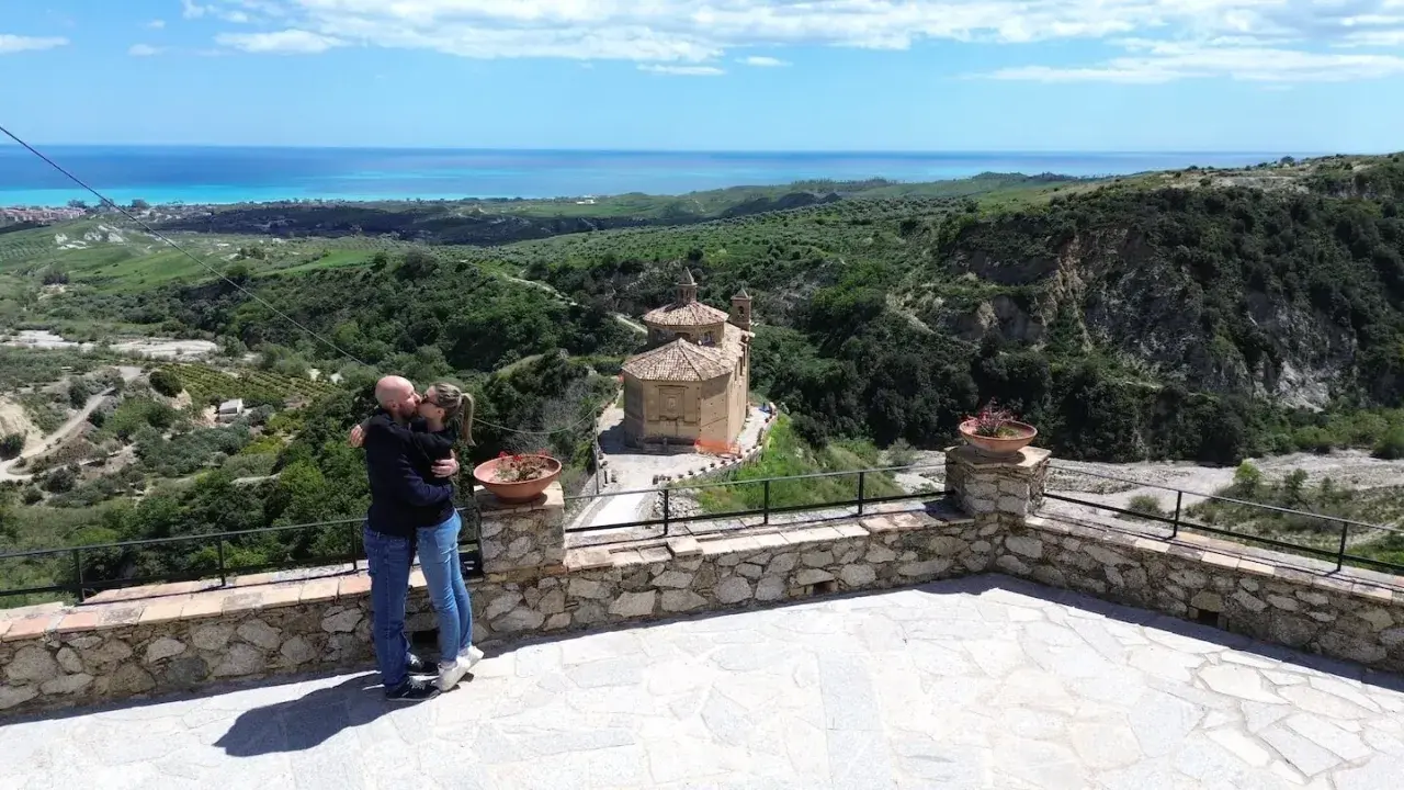 Coppia si bacia con vista panoramica su Badolato, Calabria, con il mare all'orizzonte e una chiesa antica.