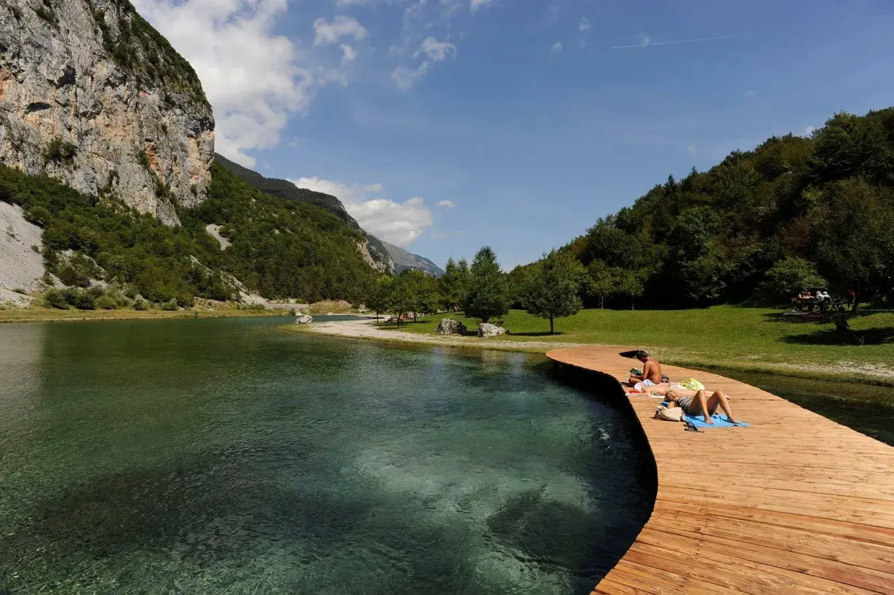 Persone si rilassano sul pontile in legno del lago di Nembia, circondato da montagne verdi e acqua cristallina.