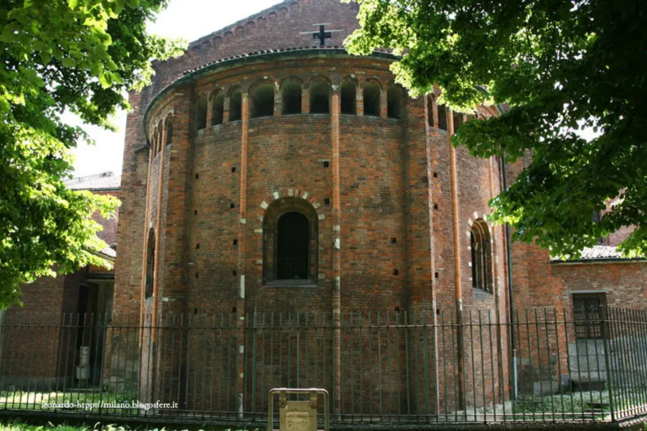 Abside in mattoni di San Nazaro in Brolo, con archetti pensili e una finestra ad arco. Alberi verdi incorniciano la scena.