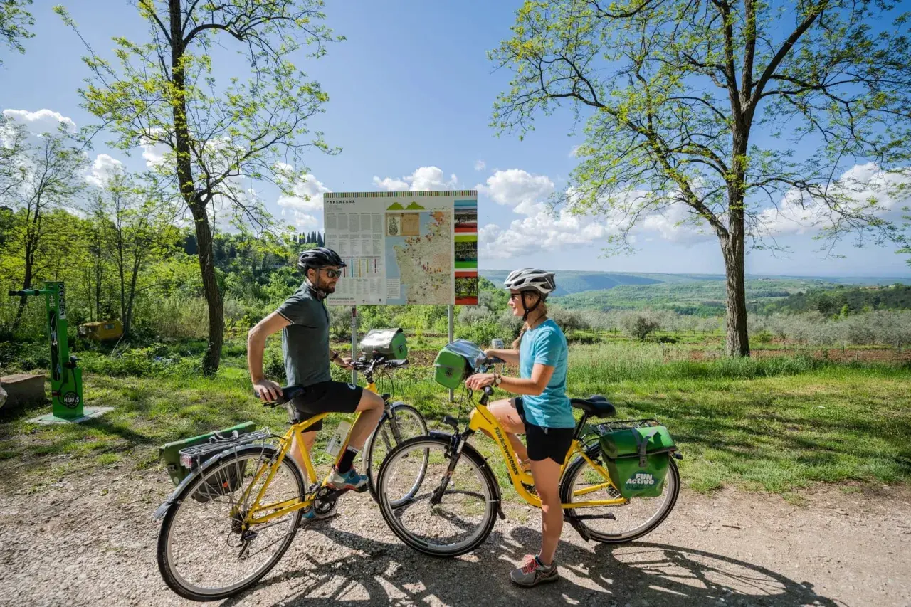Coppia in bicicletta sulla ciclabile Parenzana, con mappa del percorso e paesaggio collinare sullo sfondo.