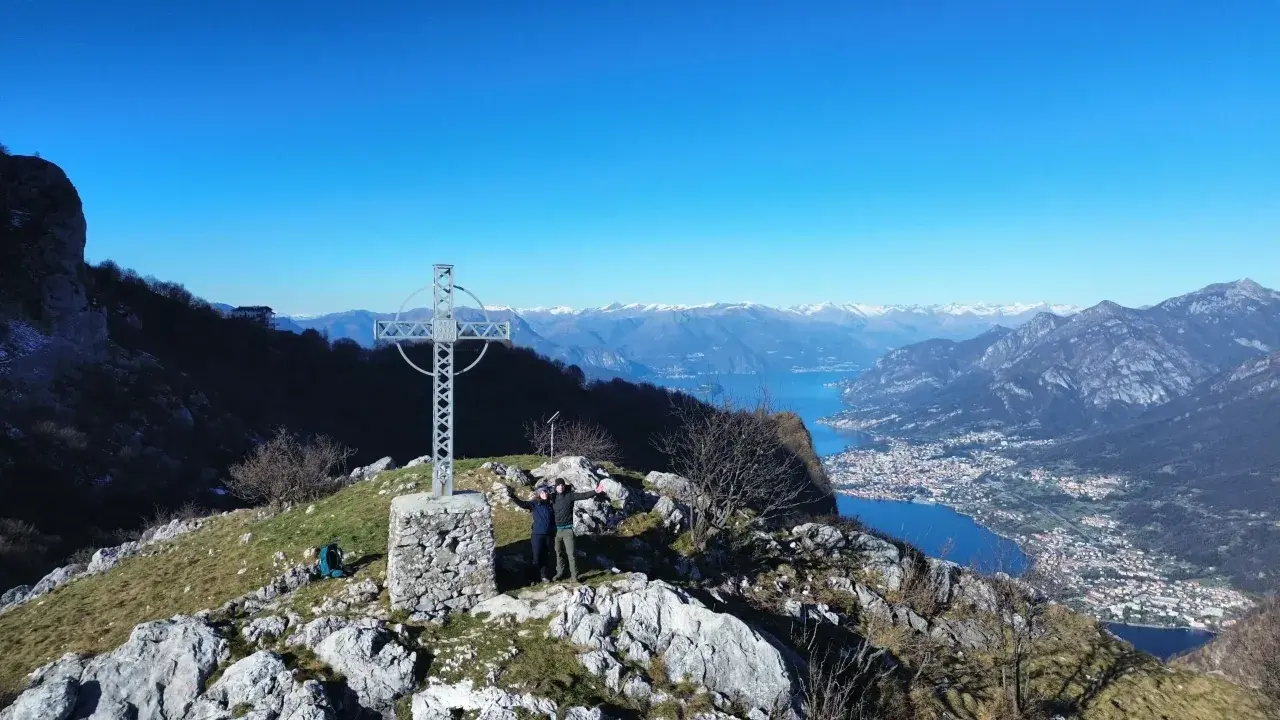Due escursionisti celebrano la vetta dei Corni di Canzo con una croce e un panorama mozzafiato sul lago.