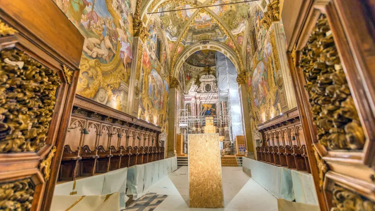 Interno della Certosa di Pavia, con affreschi dorati, stalli lignei e un'impalcatura. Cosa vedere in questo capolavoro.