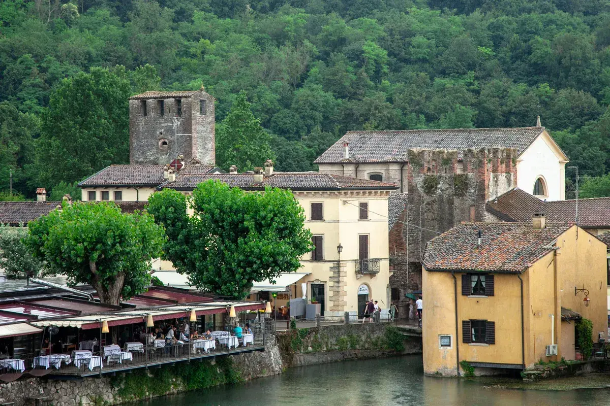 Un pittoresco borghetto sul Mincio con case storiche, torri medievali e un ristorante con tavoli all'aperto affacciato sul fiume.