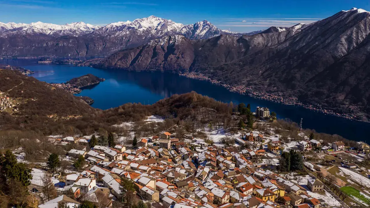 Paesaggio invernale nel Val d'Intelvi con case innevate, lago blu e montagne maestose sullo sfondo.