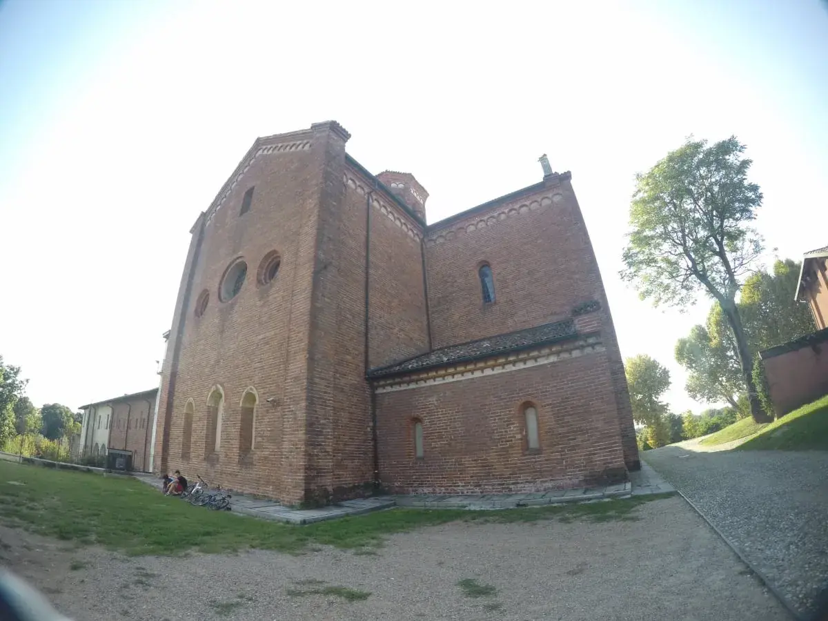 L'imponente facciata in mattoni dell'abbazia di Morimondo si staglia contro il cielo. Alcune persone e biciclette riposano sull'erba.
