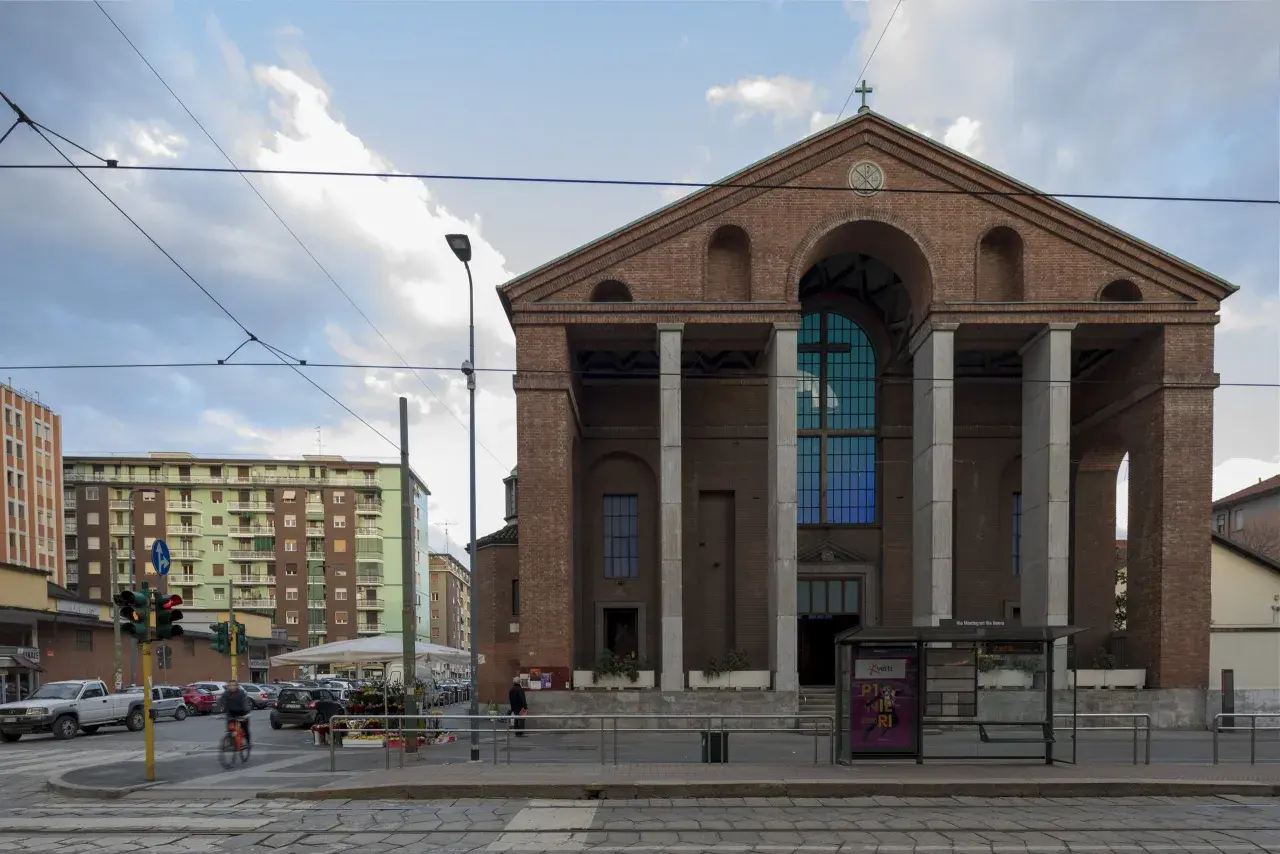 La facciata in mattoni rossi di una chiesa a Milano, con un grande rosone blu e colonne imponenti. Sullo sfondo, palazzi residenziali e tram.