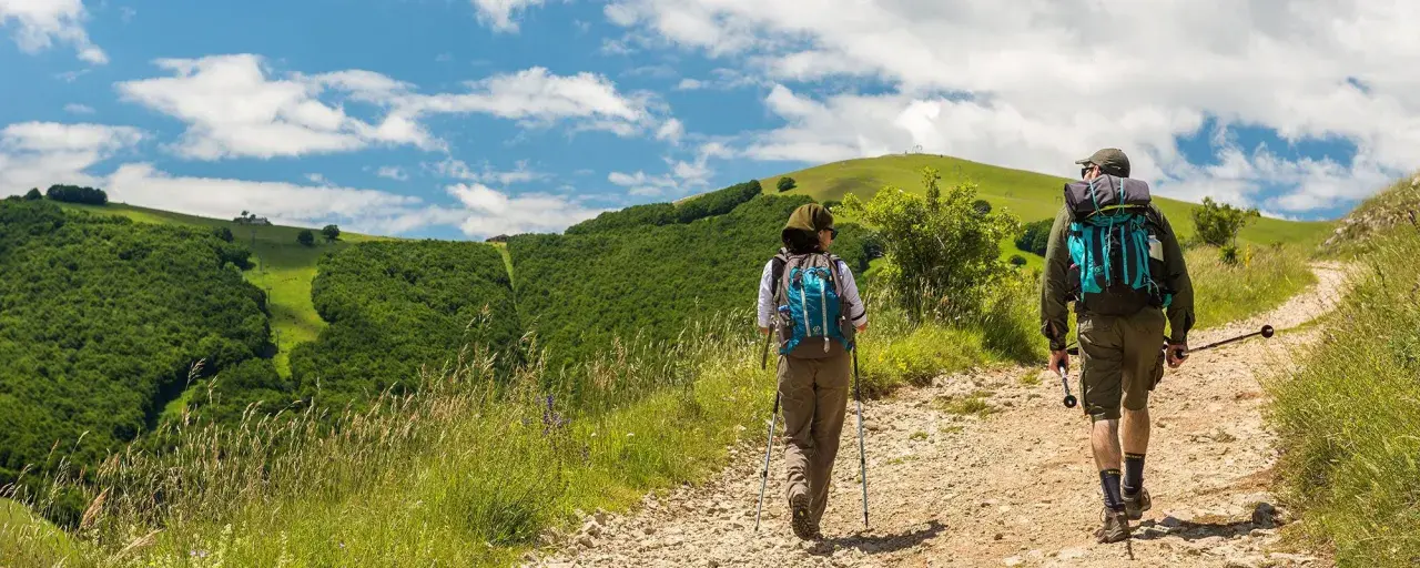 Due escursionisti affrontano un sentiero collinare, con lo sguardo rivolto verso l'orizzonte. La distanza tra Assisi e Loreto sembra un viaggio spirituale e fisico.