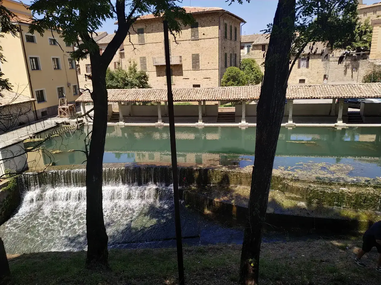 Cascata e portico storico a Bevagna, Umbria. L'acqua scende su gradoni di pietra, riflettendo gli edifici antichi.