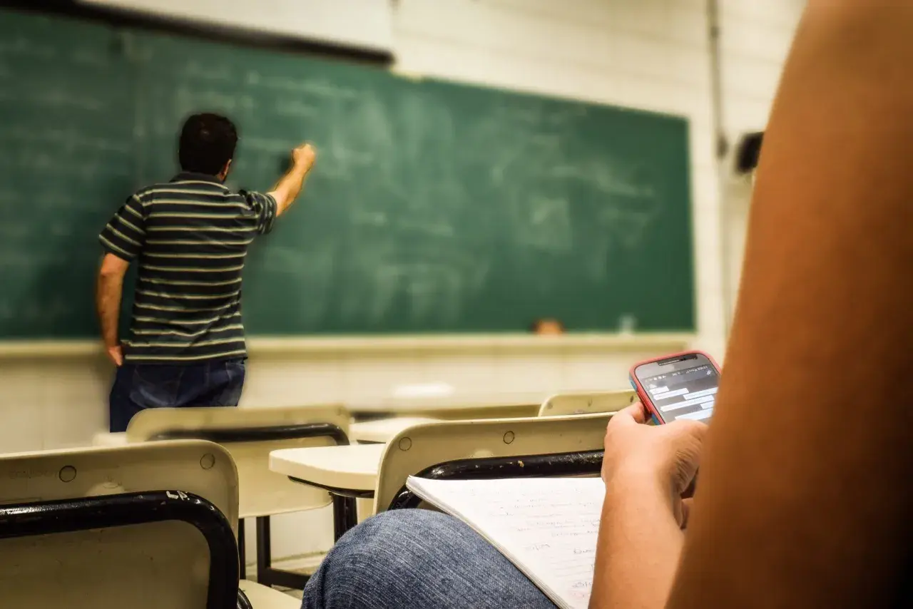 Studente distratto al cellulare mentre il professore scrive alla lavagna, forse un nuovo concorso scuola in arrivo.