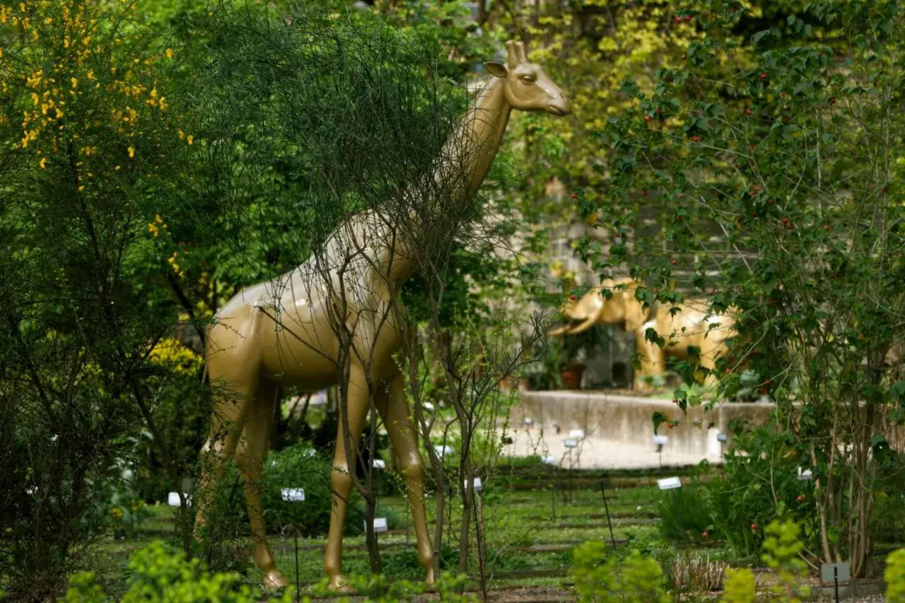 Una giraffa dorata emerge tra la vegetazione lussureggiante dell'orto botanico di Brera, con un elefante dorato sullo sfondo.