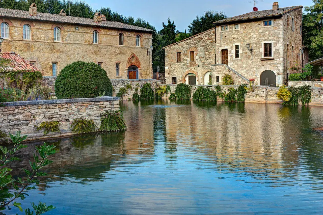 Antiche terme libere a San Gimignano, con edifici in pietra che si specchiano in un laghetto tranquillo.