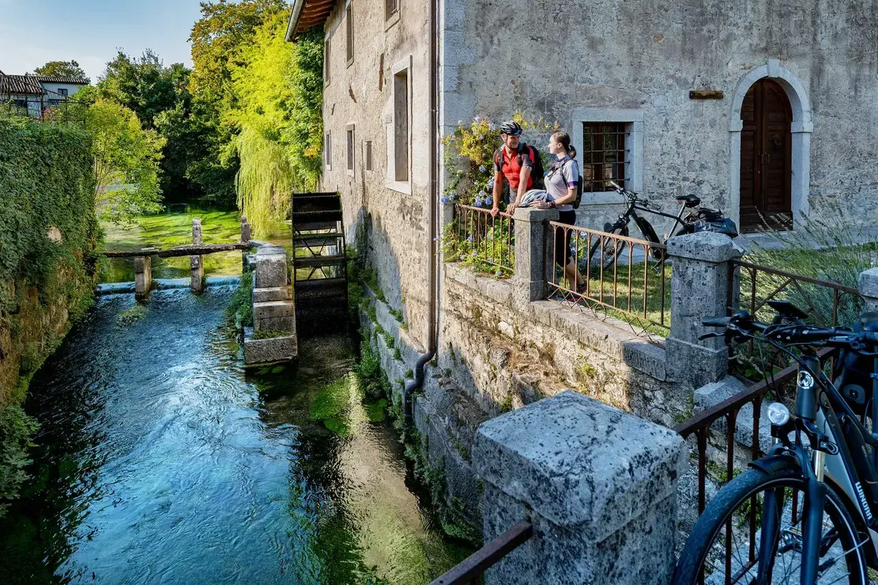 Coppia in bicicletta ammira un antico mulino ad acqua, uno dei tanti borghi friulani da visitare, immersi nel verde e lungo un ruscello cristallino.
