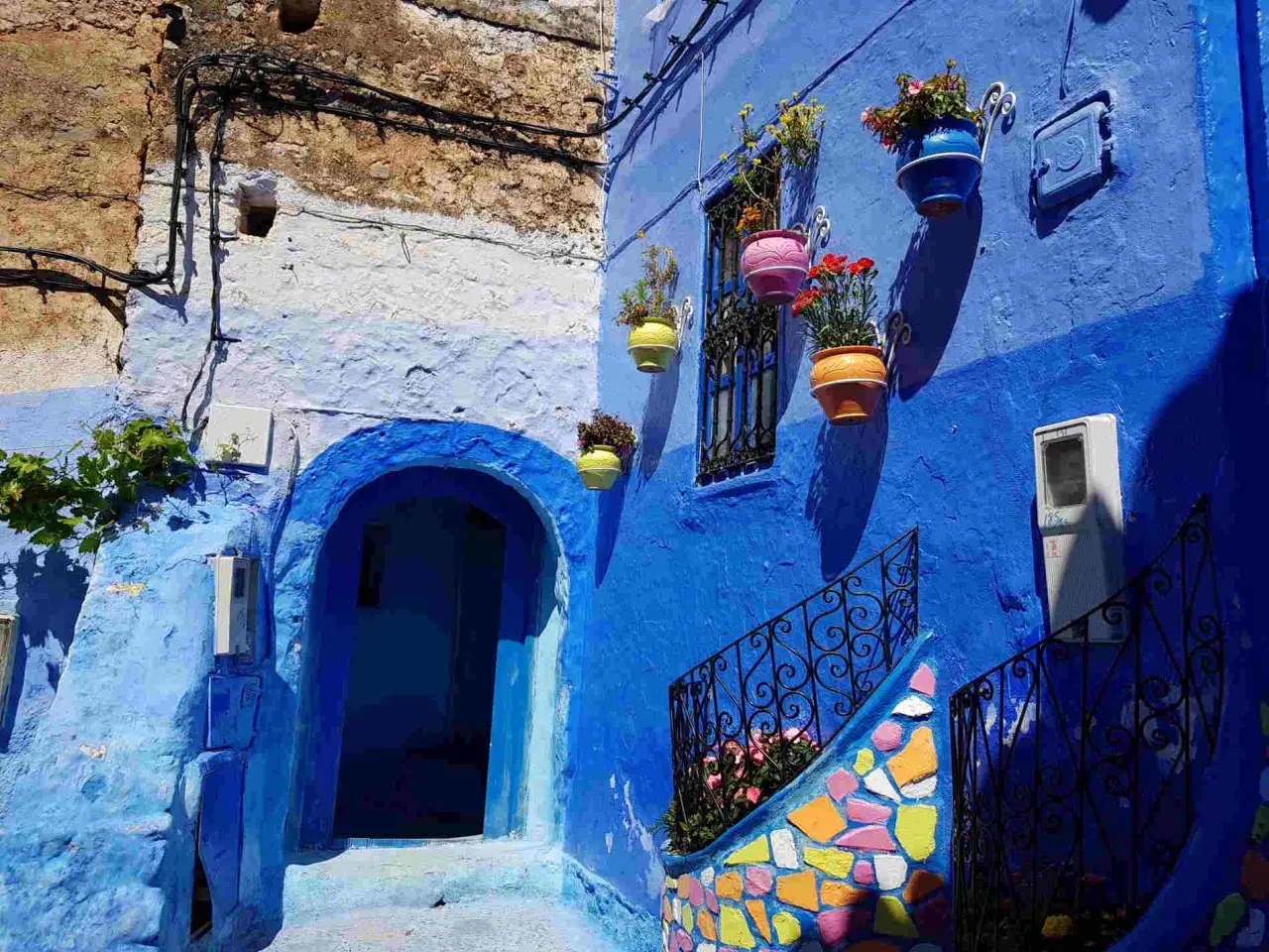 Un vicolo nella città azzurra del Marocco, con case blu, vasi di fiori colorati e una scala decorata.