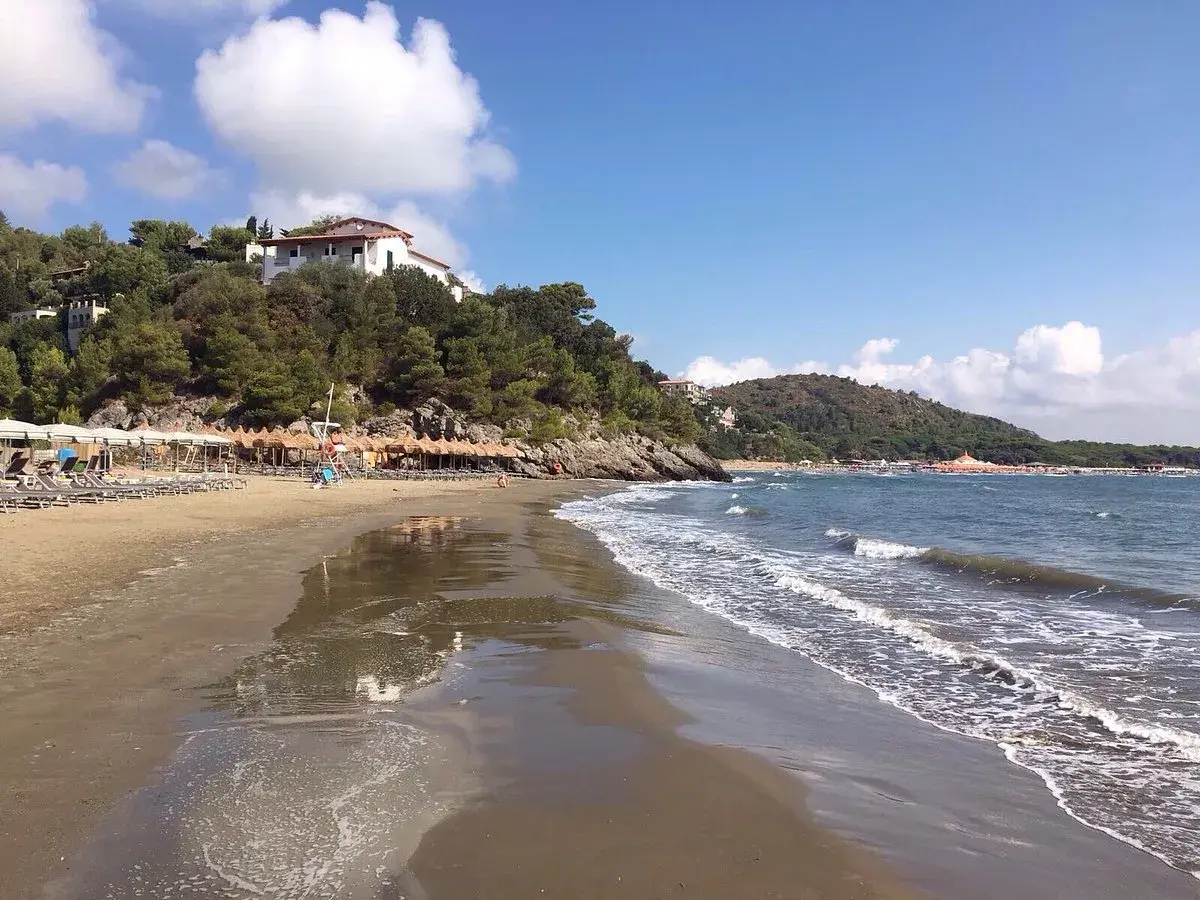 Spiaggia di **Porto d'Ercole** con ombrelloni, lettini e mare calmo sotto un cielo azzurro con nuvole bianche.