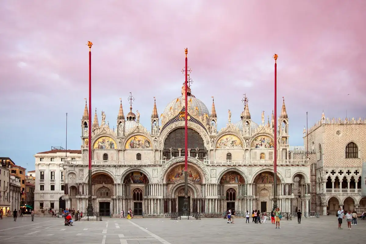 La maestosa chiesa San Marco a Venezia, con le sue cupole e mosaici, si staglia contro un cielo rosa al tramonto.