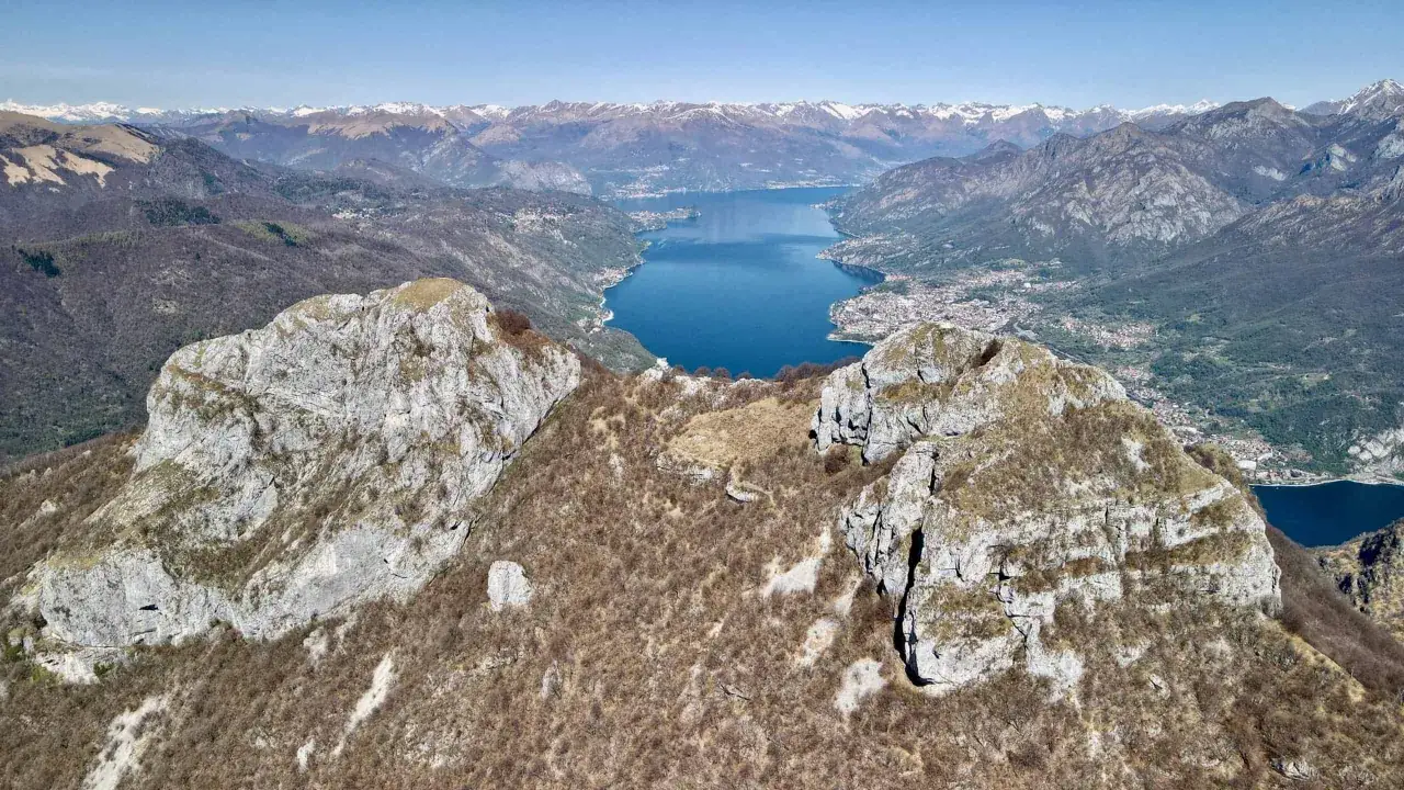 Vette rocciose dei Corni di Canzo dominano un lago azzurro e montagne innevate all'orizzonte.