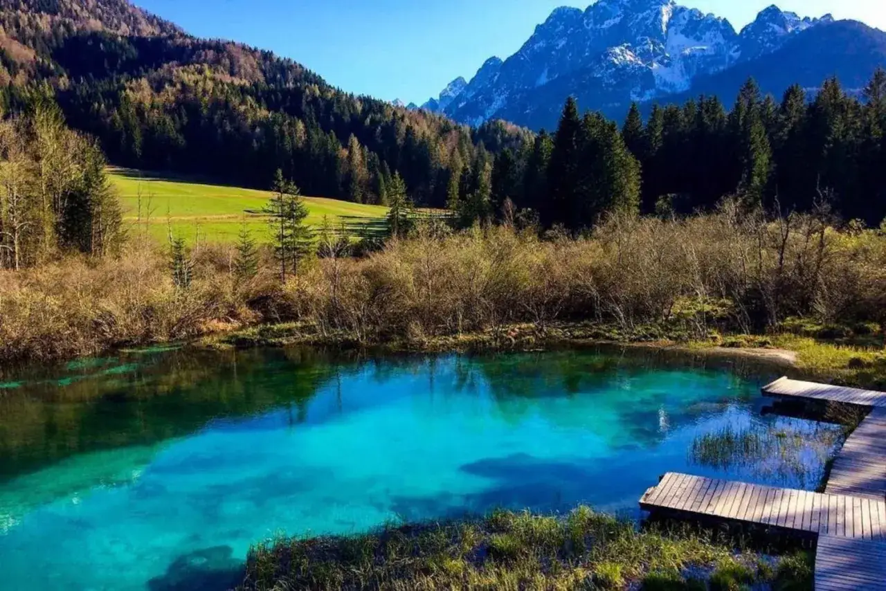 Sorgente cristallina nel Parco Nazionale del Tricorno, con montagne innevate sullo sfondo e un pontile in legno.