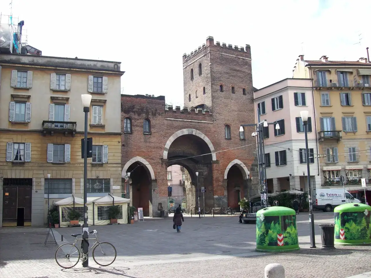 La Porta Ticinese, con la sua imponente struttura in mattoni, domina la piazza. Edifici colorati la circondano, mentre una persona cammina verso l'arco.