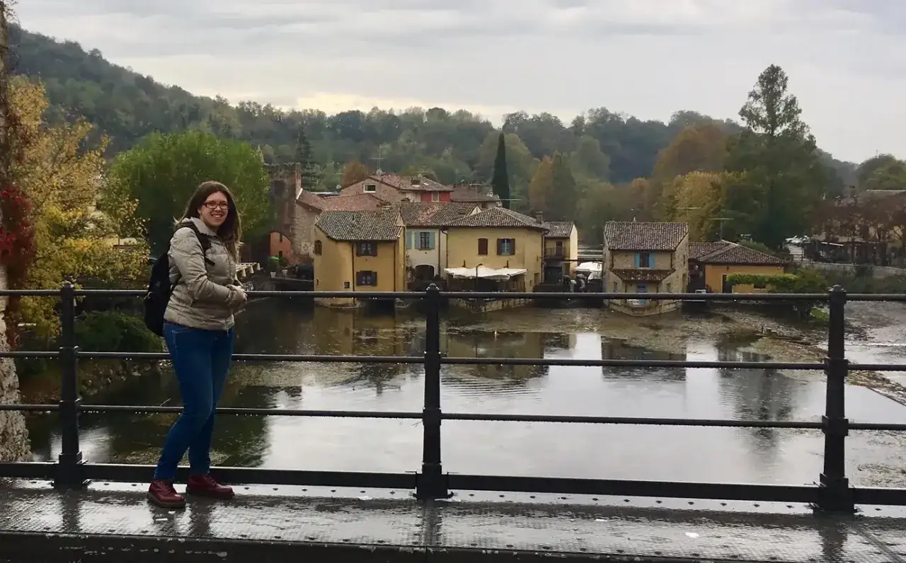 Una ragazza sorride su un ponte a Borghetto sul Mincio, con case pittoresche sull'acqua e alberi autunnali sullo sfondo.