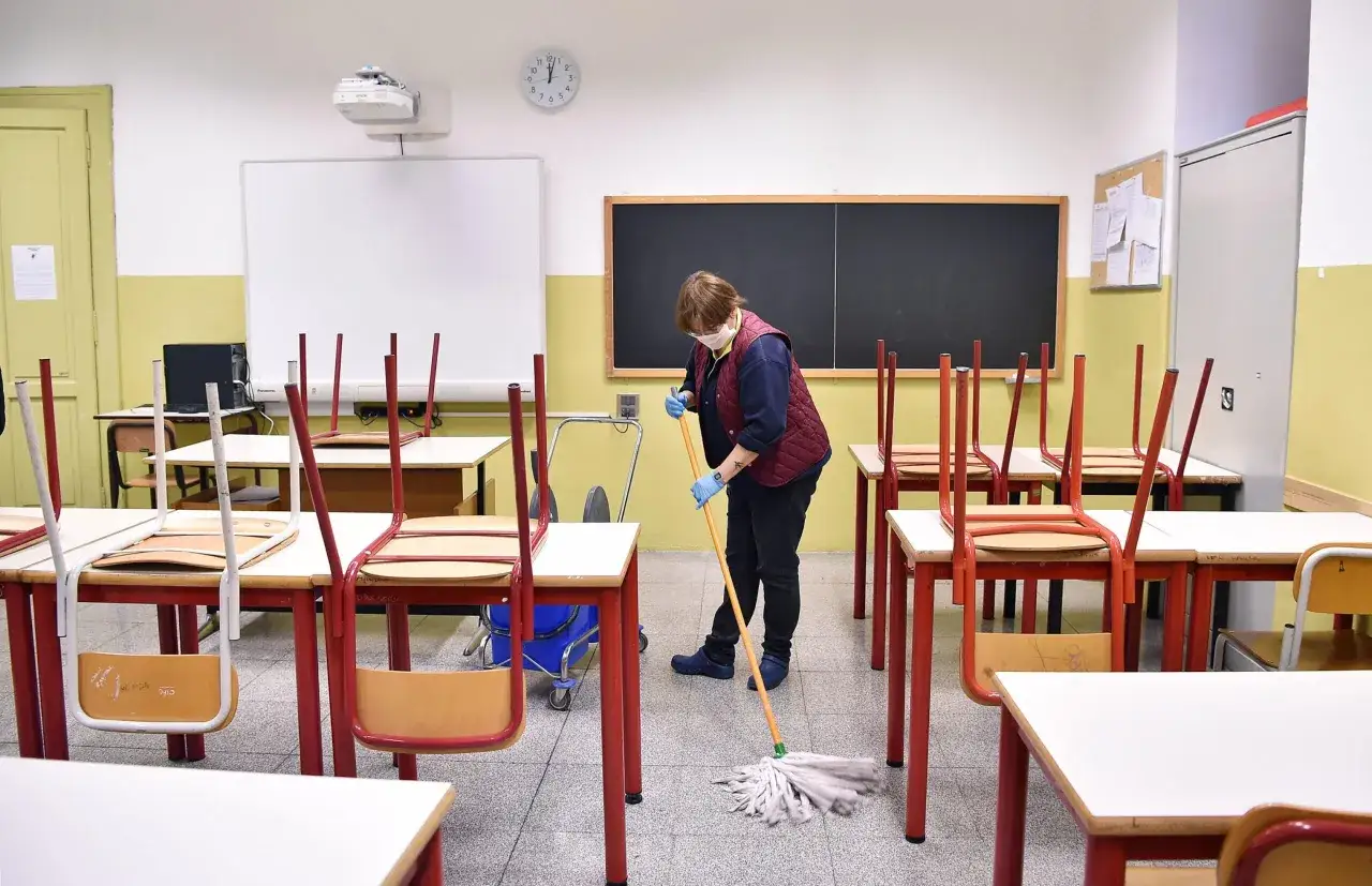 Un collaboratore scolastico pulisce l'aula con uno straccio. Le sedie sono rovesciate sui banchi.
