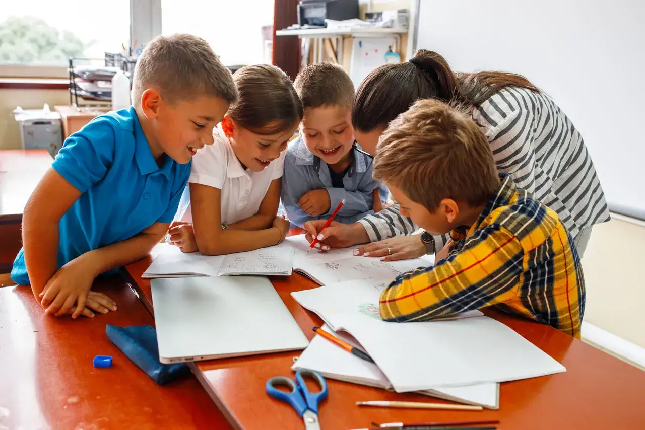 Bambini e insegnante collaborano a un progetto di scuola sostegno, sorridendo mentre scrivono e disegnano su quaderni.