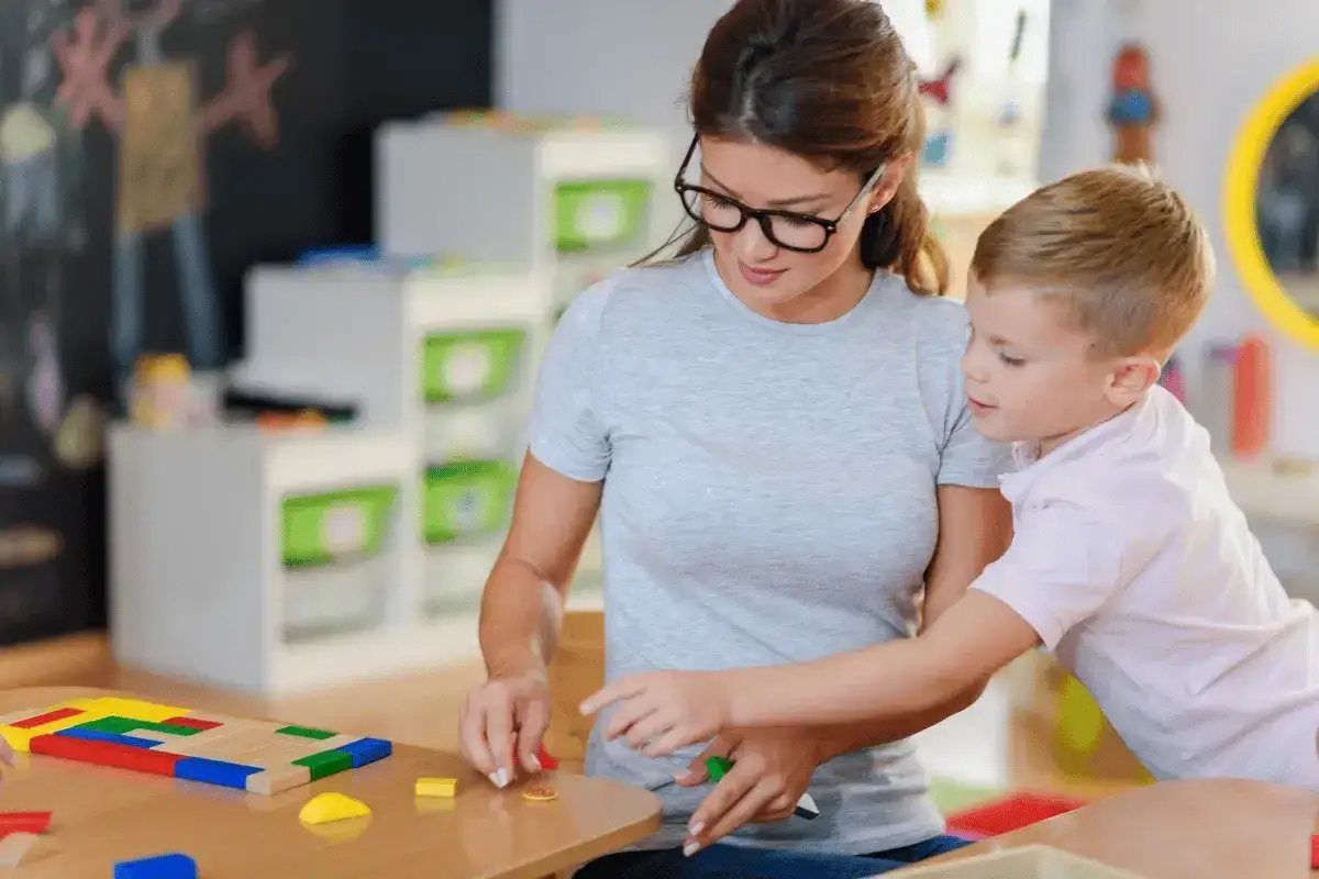 Donna con occhiali aiuta bambino con blocchi colorati. Un esempio di motivazione TFA sostegno orale, apprendimento ludico.
