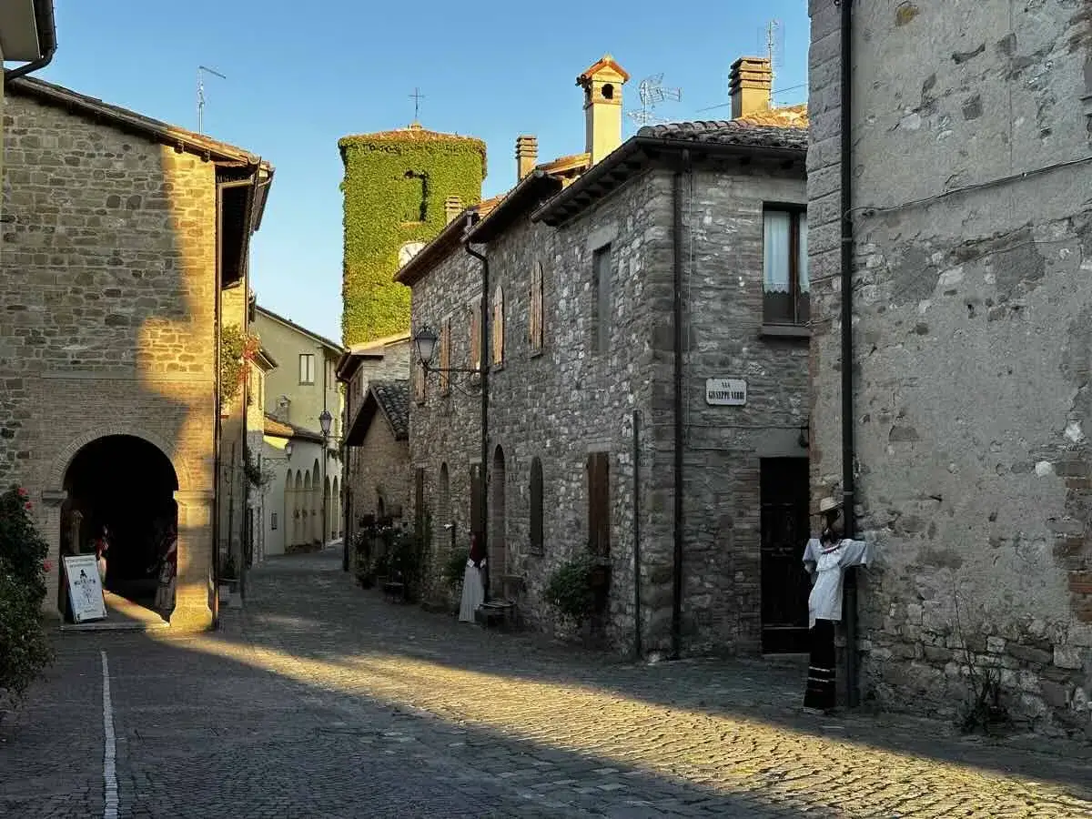 Un vicolo acciottolato a Macerata Feltria, con case in pietra e una torre verdeggiante.