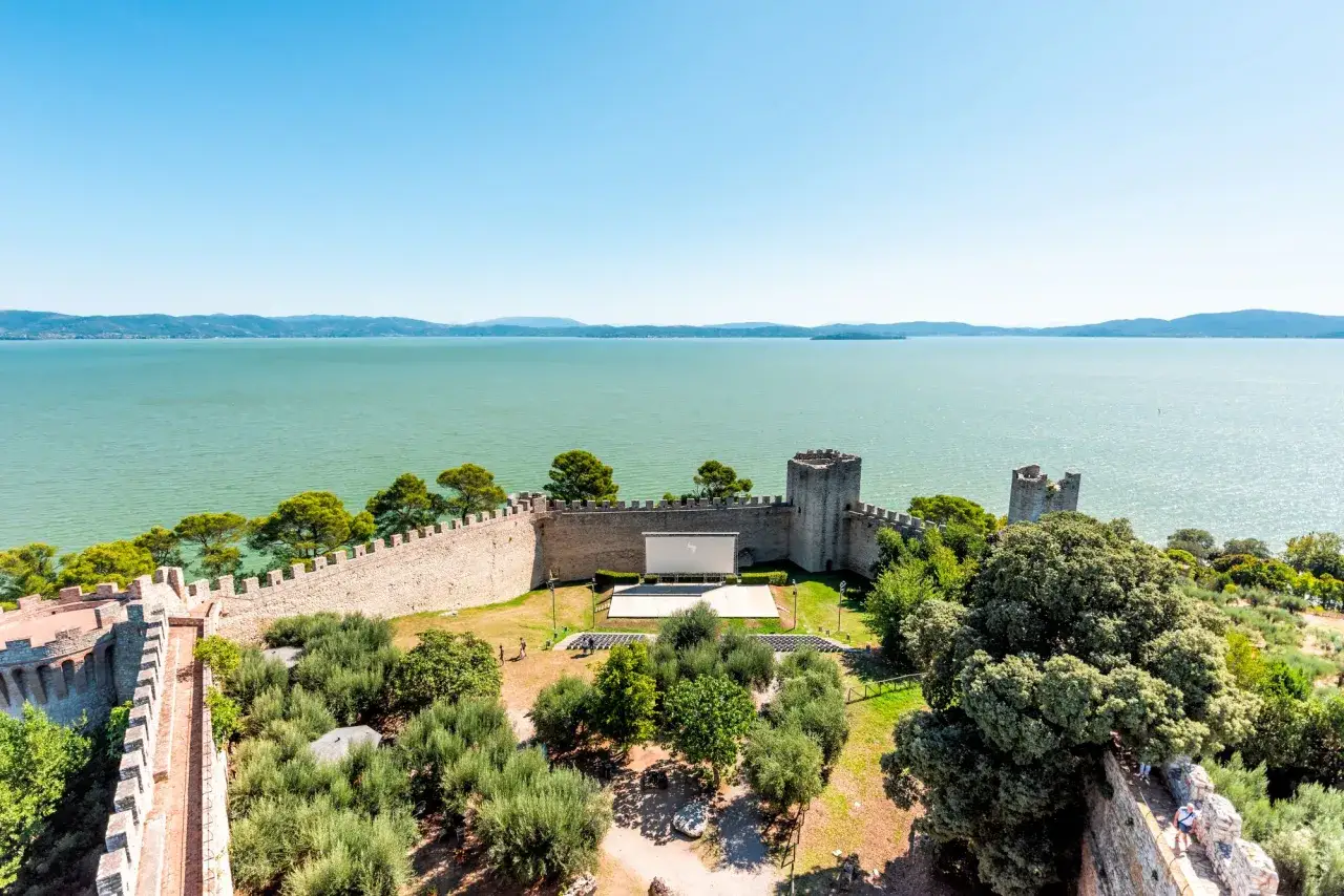 Vista aerea del lago e delle mura di Castiglione del Lago. All'interno, un cinema all'aperto con schermo e sedute.