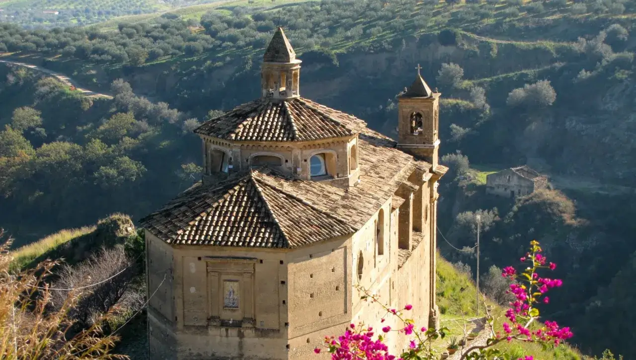 Chiesa antica su una collina a Badolato, Calabria, con uliveti e un rudere sullo sfondo.