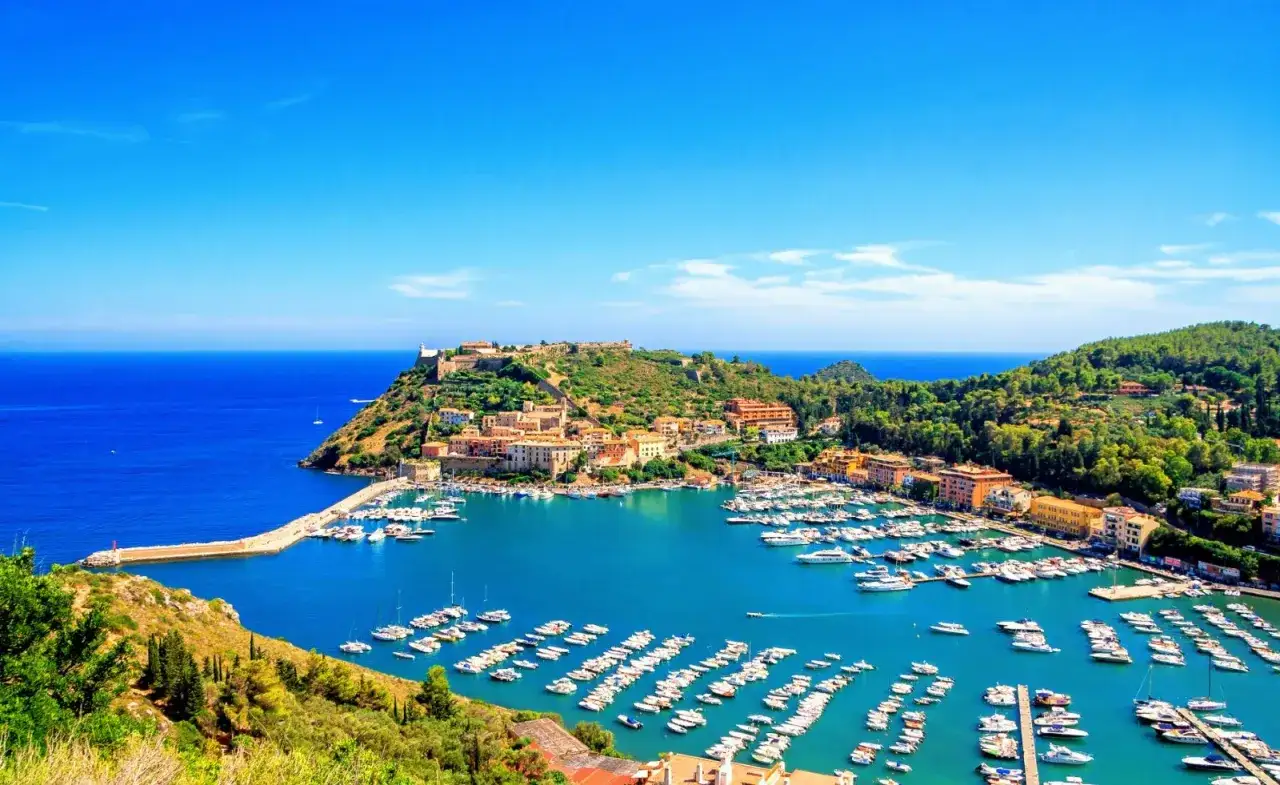 Vista panoramica del porto d'Ercole con il mare blu, le barche ormeggiate e le case colorate che si affacciano sulla costa.