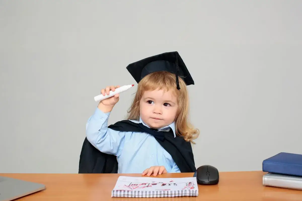 Bambino con tocco e toga, pronto per l'esame di quinta elementare, tiene un pennarello bianco.