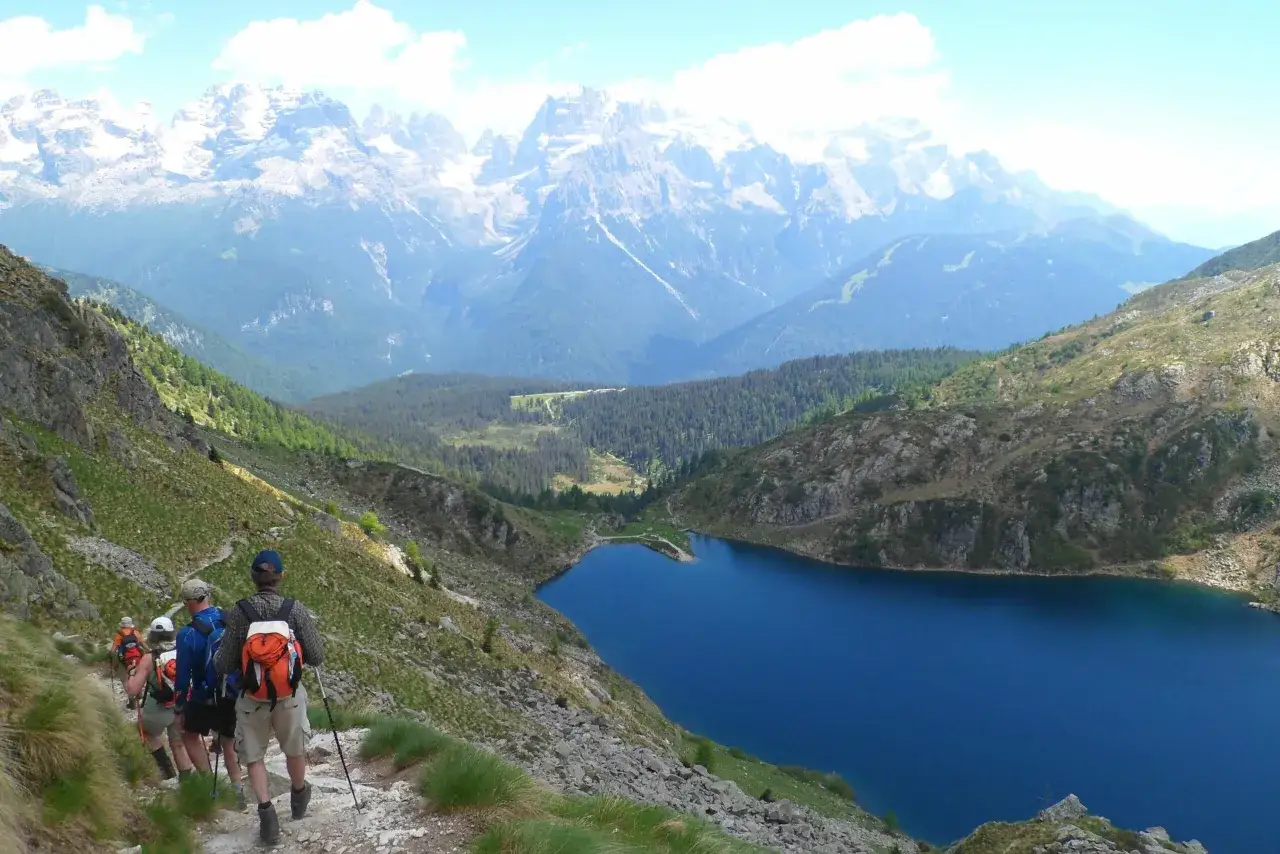 Escursionisti sul sentiero del giro dei 5 laghi a Madonna di Campiglio, con un lago blu intenso e le maestose Dolomiti sullo sfondo.