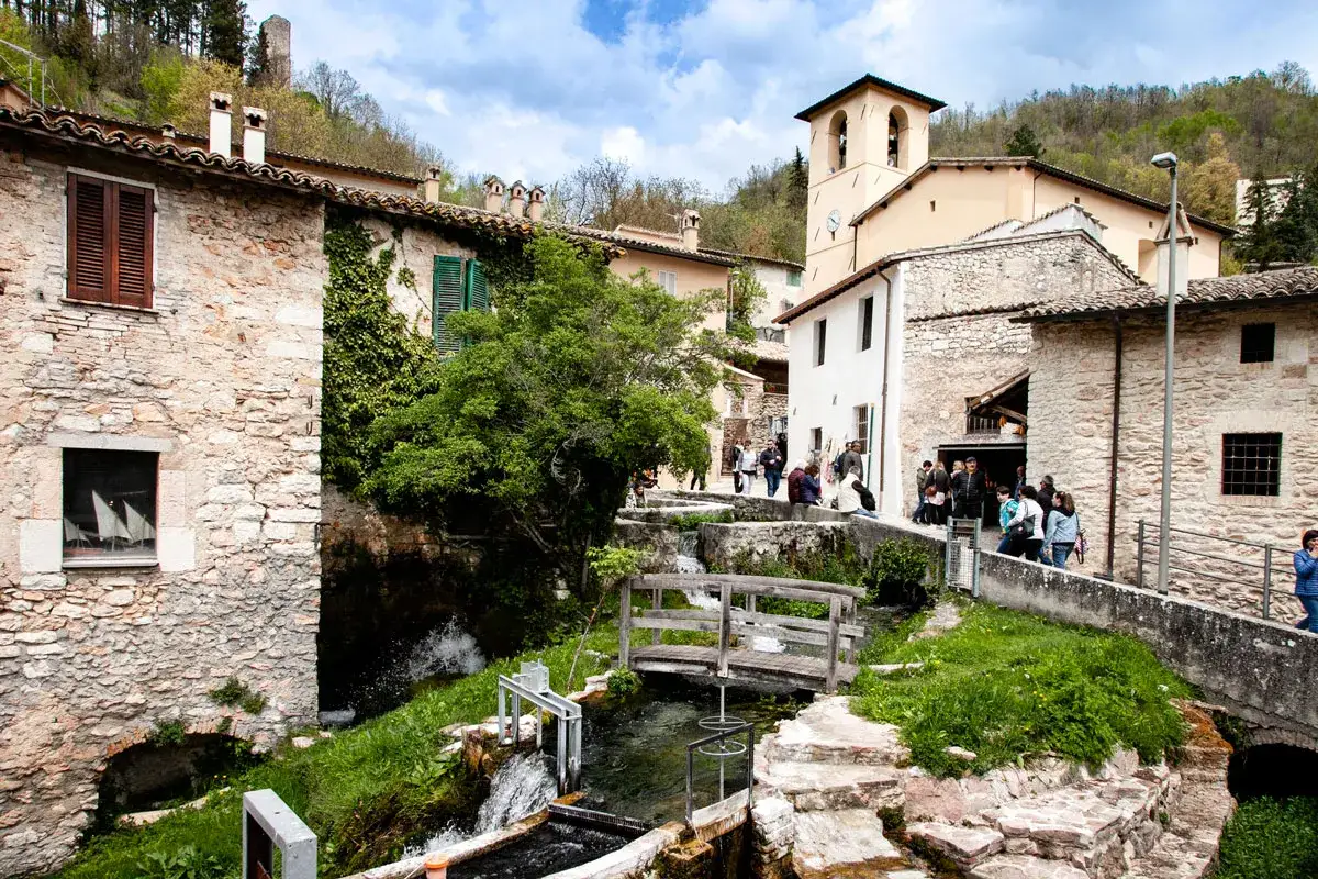 Un borgo in Umbria con cascate d'acqua che scorrono tra antiche case di pietra. Un ponte di legno attraversa il ruscello, mentre turisti passeggiano tra le vie.