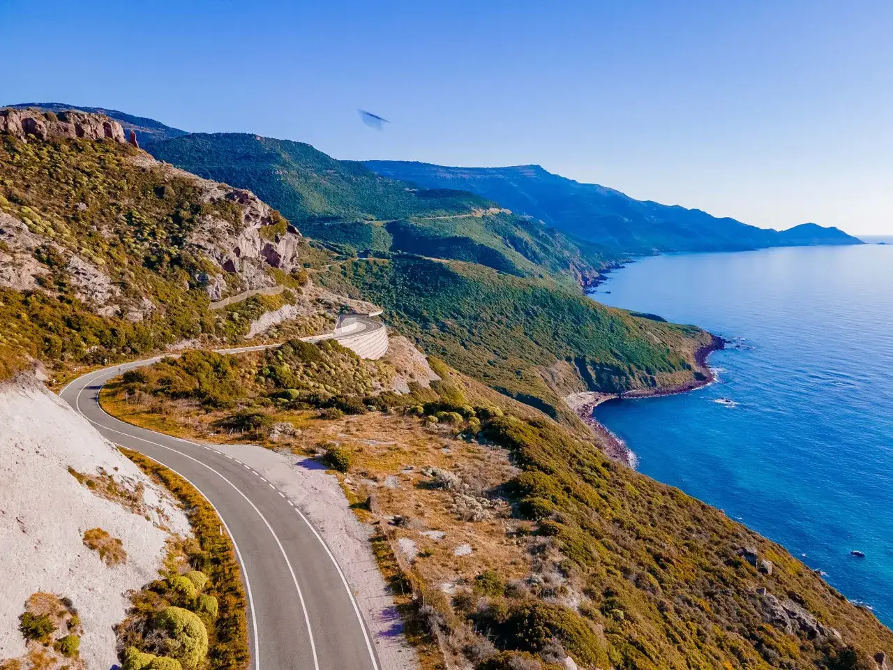 Strada panoramica lungo la costa tra Alghero e Bosa, con mare blu intenso e montagne verdi.