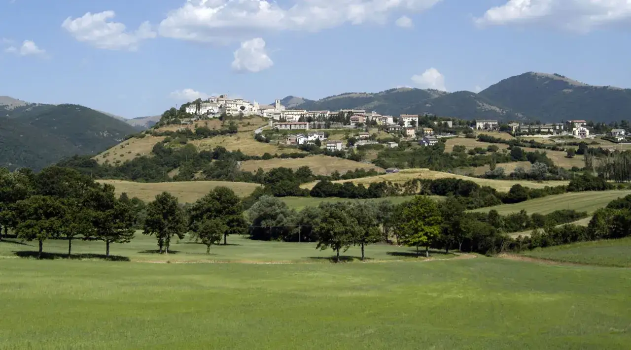 Panoramica di Monteleone di Spoleto, un borgo arroccato su una collina, circondato da verdi colline e campi coltivati sotto un cielo azzurro con nuvole bianche.