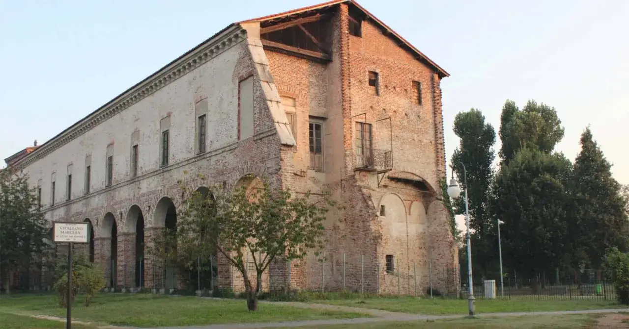 Il castello di Melegnano, con i suoi mattoni a vista e le arcate, si staglia contro il cielo sereno, circondato da alberi verdi.
