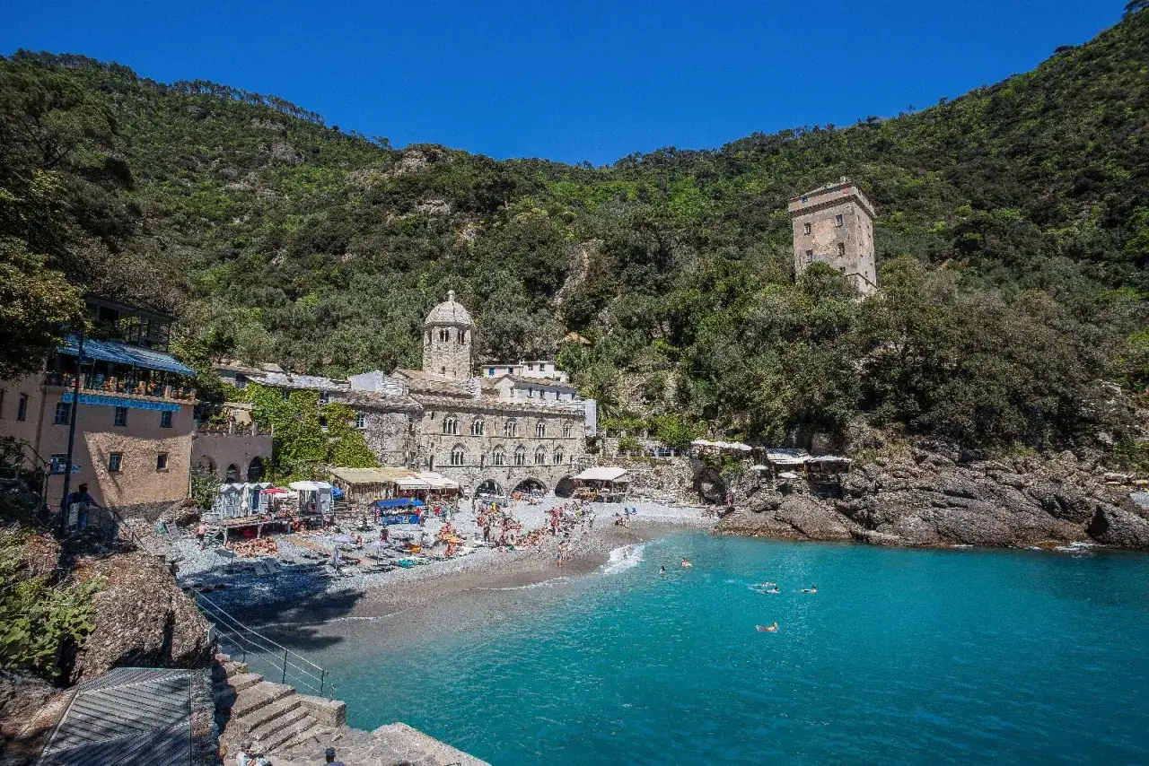 Baia di San Fruttuoso, Portofino: spiaggia affollata, mare azzurro, antica abbazia e torre medievale incastonati nel verde.